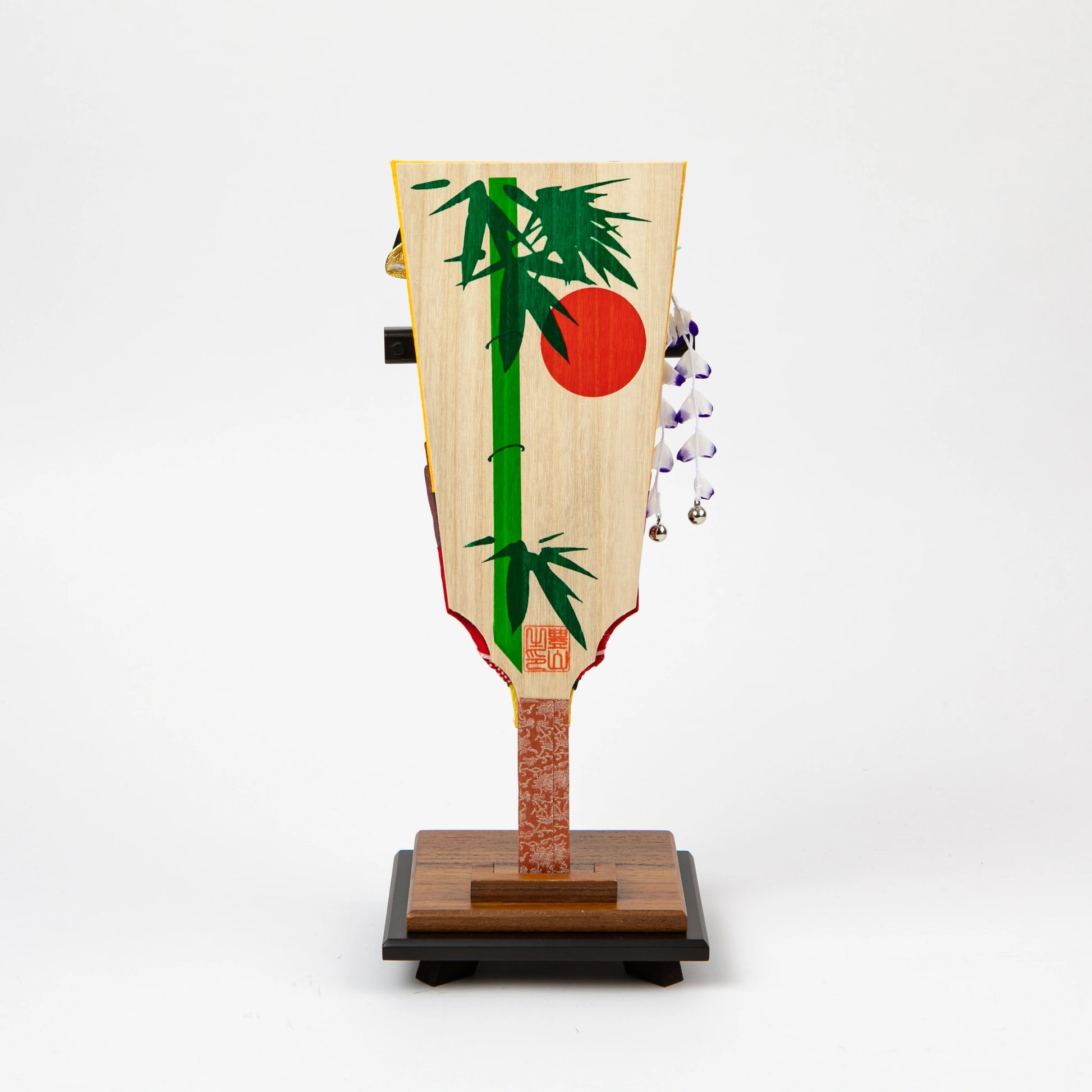 Back of a decorative hagoita paddle featuring a painted green bamboo and red sun motif on a natural wood background, displayed on a wooden stand.