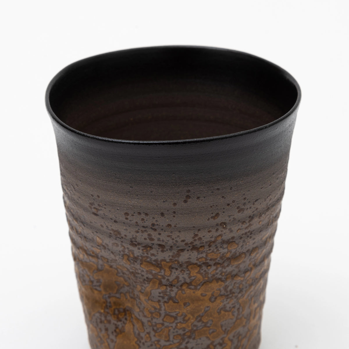 A close up of the upper part of a handleless ceramic tumbler featuring a dark brown ribbed upper section and a heavily textured, speckled ochre glaze on the lower half, set against a white background.