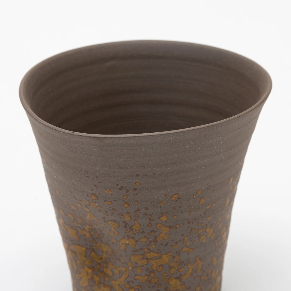 A handleless brown ceramic tumbler close up featuring a heavily textured, speckled ochre glaze on the lower half, set against a white background.