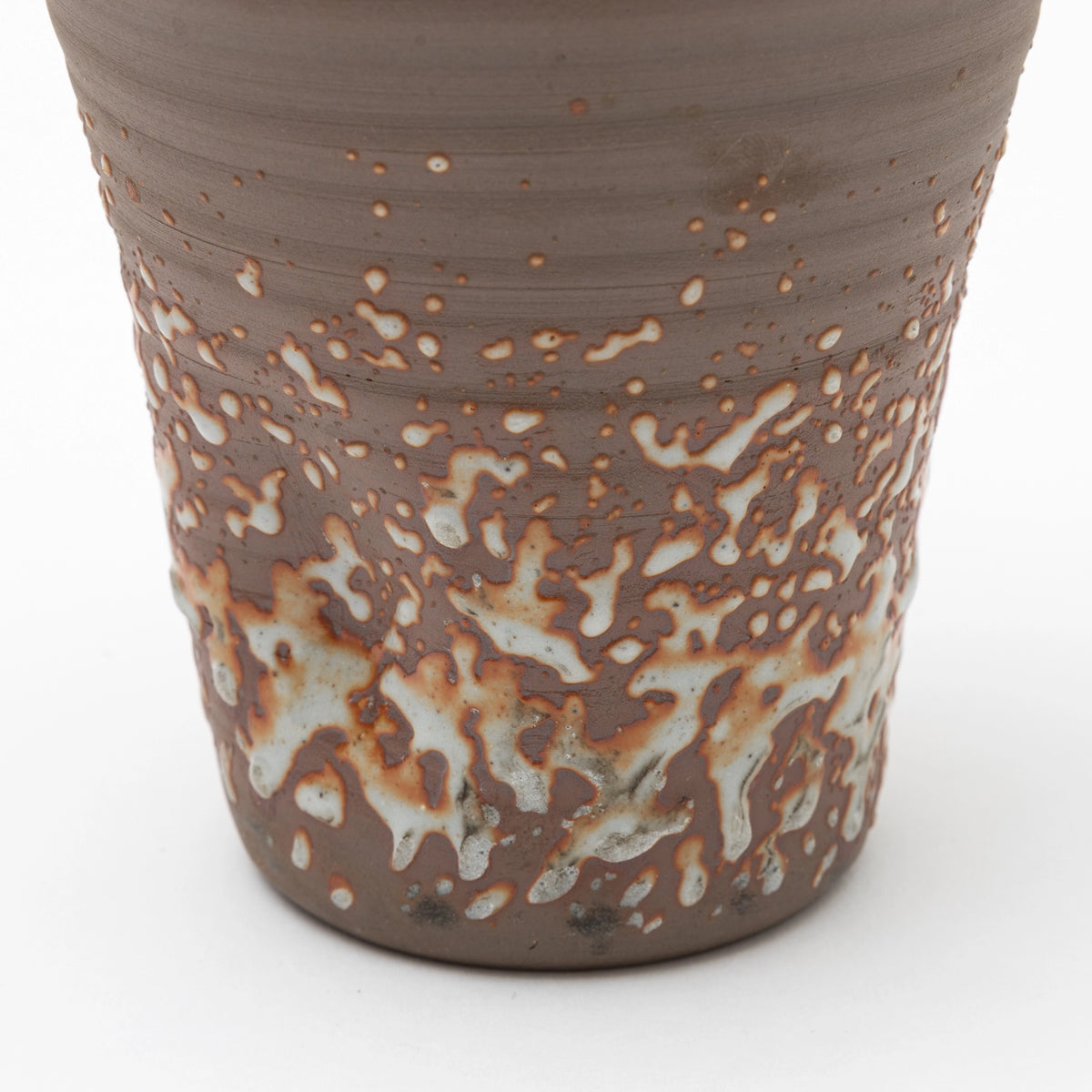 A close up of the lower part of a handleless brown ceramic tumbler featuring a heavily textured, speckled white glaze on the lower half, set against a white background.