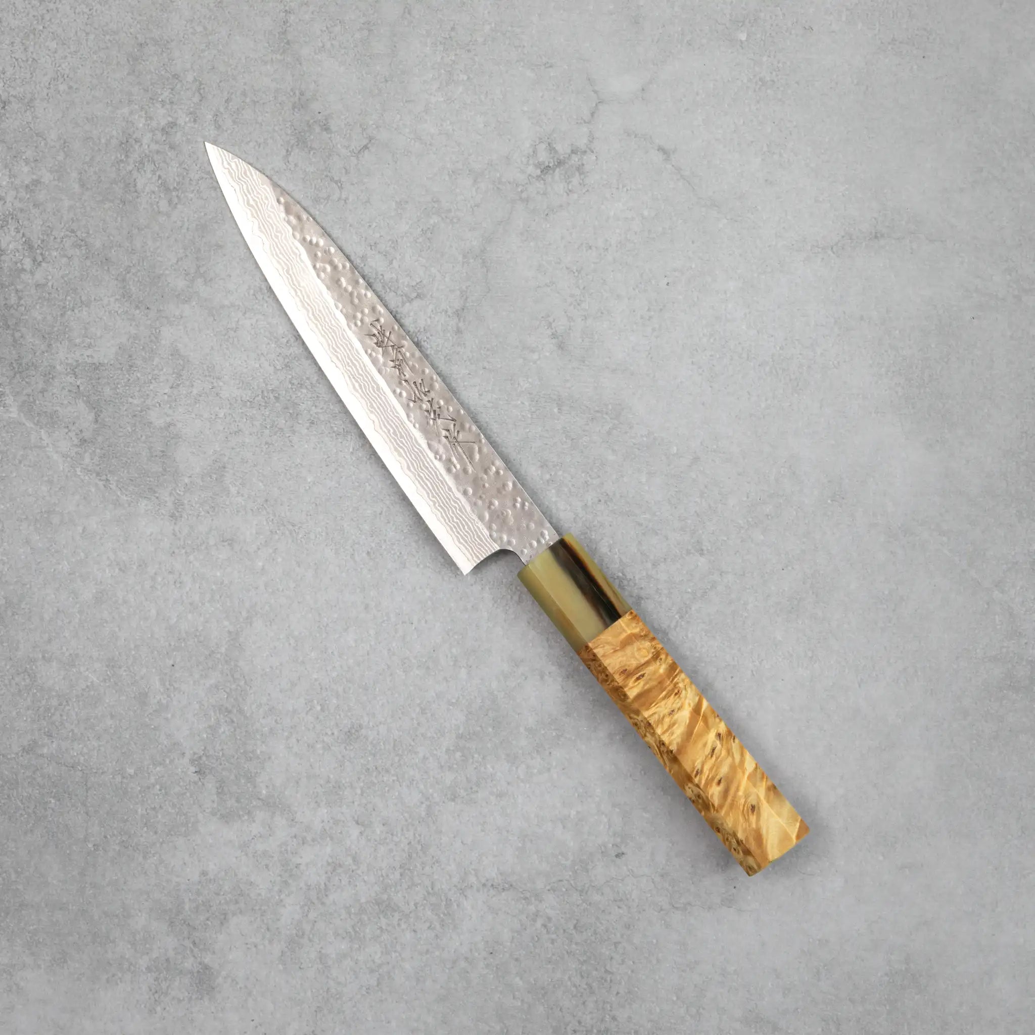 Side view of the Kamo Hamono Shizuku petty knife, showing a narrow pointed VG10 Damascus blade with rounded tsuchime hammer marks, a water buffalo horn bolster, and a stabilized burl wood handle with pronounced figured grain.