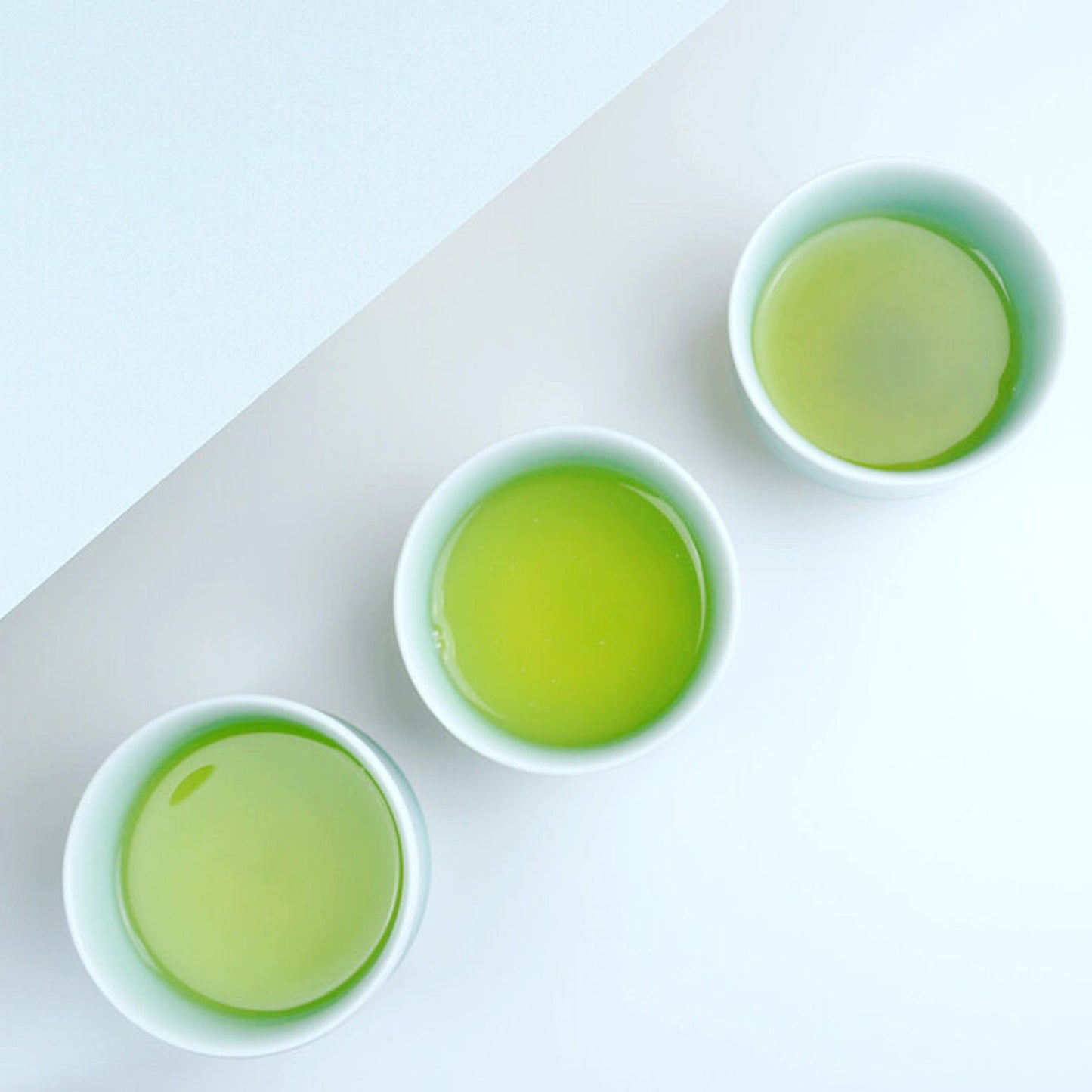 Three Nabeshima celadon porcelain tea cups filled with vibrant green tea, arranged in a row on a white surface.