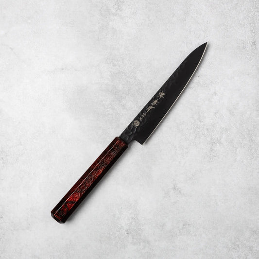 Sakai Takayuki Kurokage VG10 petty knife, featuring a hammered black blade and rich burgundy resin handle, positioned diagonally on a light stone surface.