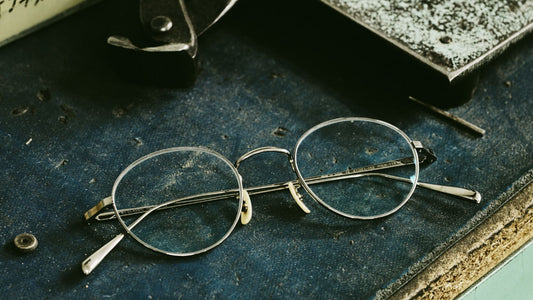 A pair of vintage round metal-frame eyeglasses lying on a rustic, blue-textured surface, surrounded by tools and dust, evoking a sense of craftsmanship and heritage.