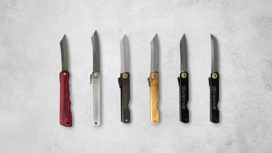 A lineup of six Higonokami knives in various handle materials and colors, including red wood, silver, black, and brass, with sharp blades and traditional Japanese engravings, displayed side by side on a light gray surface.