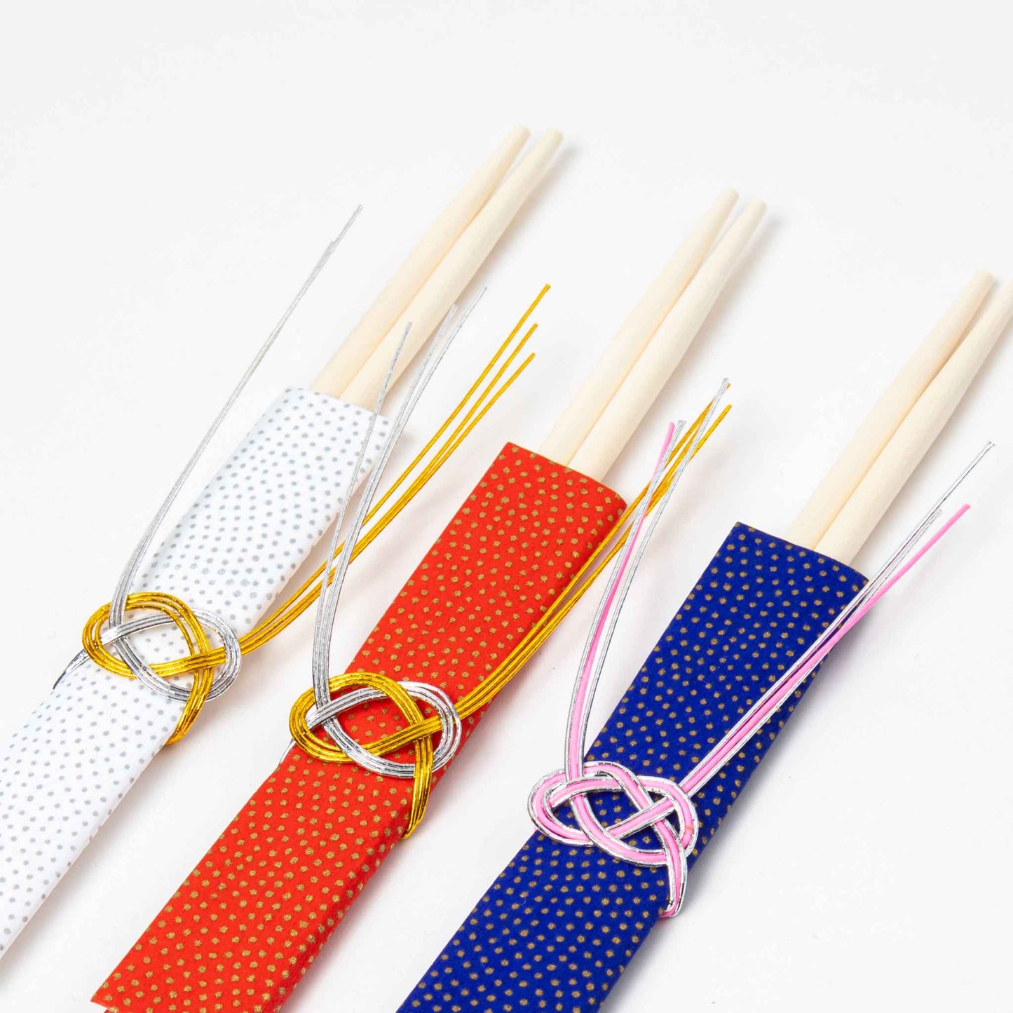 A set of three wooden chopsticks adorned with colorful dotted sleeves in white, red, and blue, each tied with intricate Mizuhiki cords in complementary metallic hues, arranged on a white background.