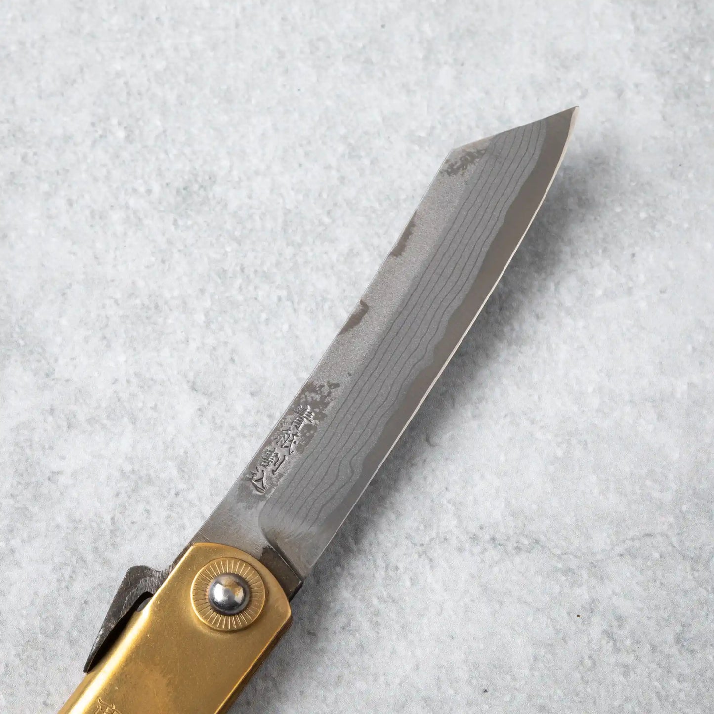 Close-up view of the Higonokami Damascus knife blade, showcasing the intricate Damascus steel pattern and engraved Japanese characters on the blade near the brass pivot.