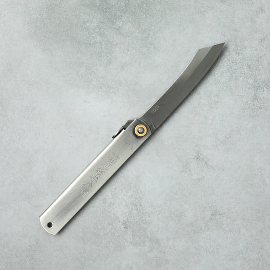 Full view of a Higonokami VG10 stainless steel knife with a silver handle and brass pivot, laid flat on a gray surface.