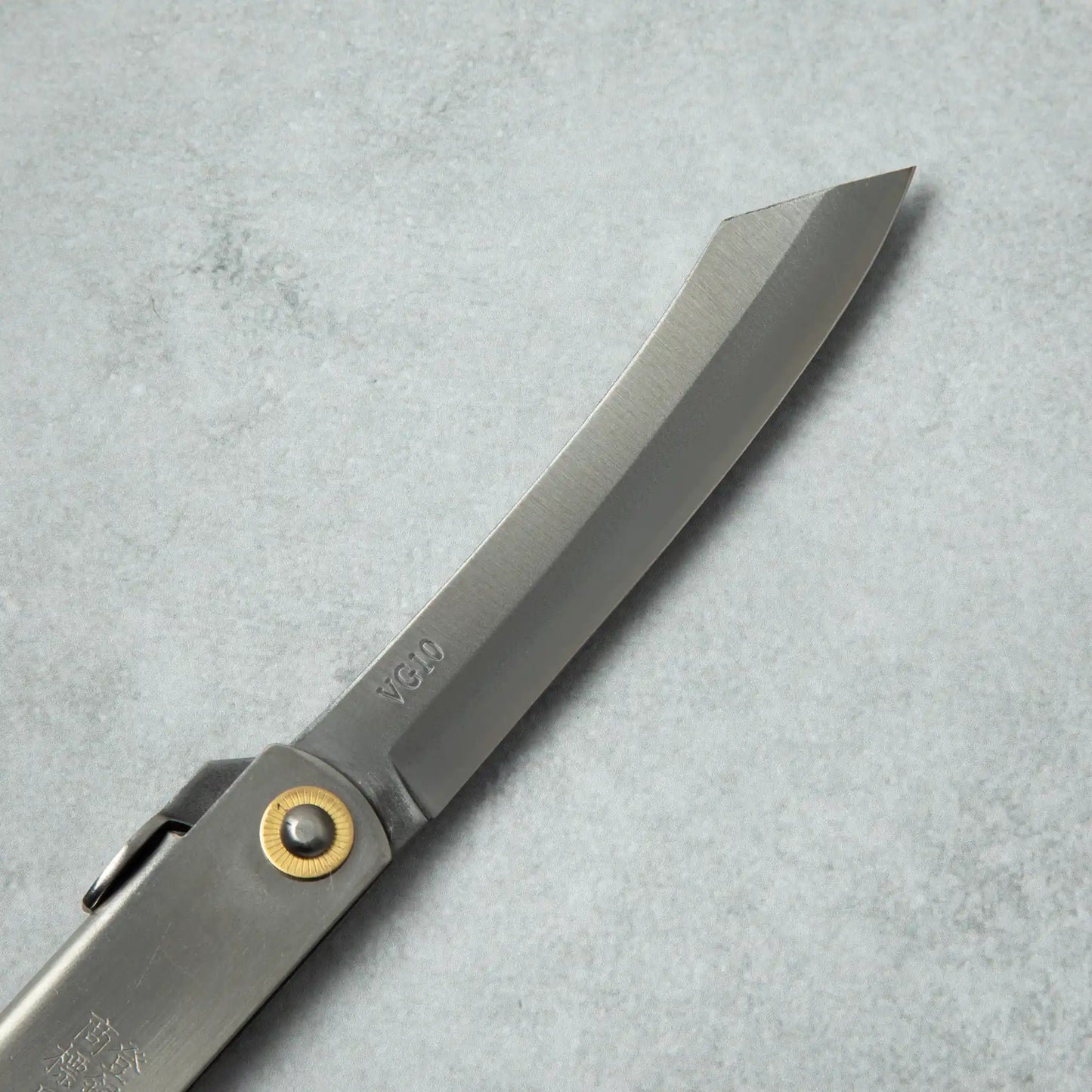 Close-up of the blade of a Higonokami VG10 stainless steel knife, featuring a curved edge and the VG10 marking near the brass pivot.