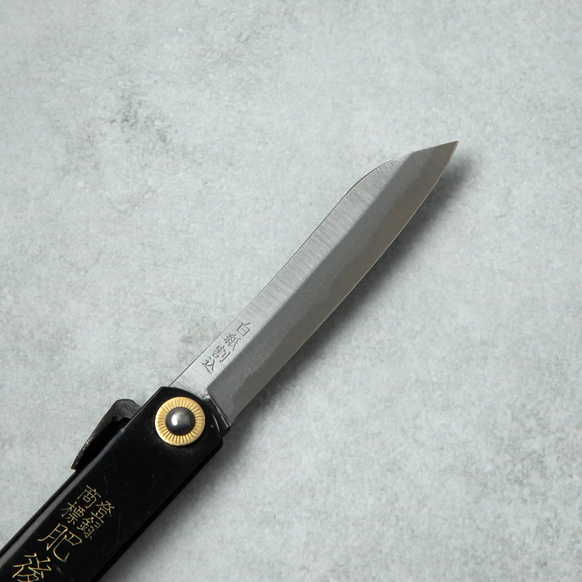 Side view of the Higonokami White Steel Bamboo knife blade, showcasing its sharp edge and brass pivot attached to a black handle.