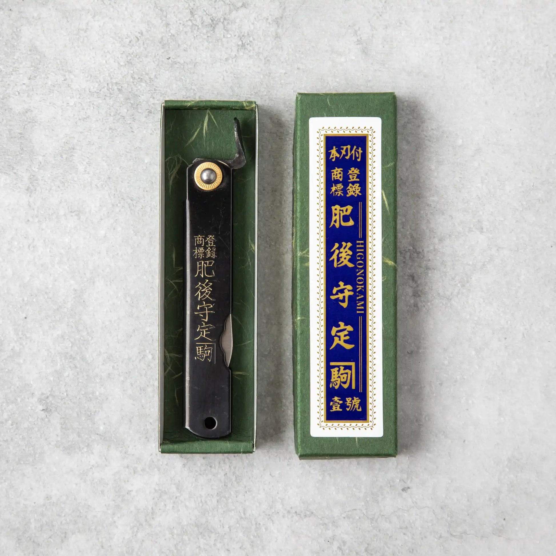 This image features the Higonokami knife with its signature Japanese script engraved on the blade. It is packaged in a traditional green box with a textured surface and gold and blue label detailing, giving it a classic Japanese aesthetic.