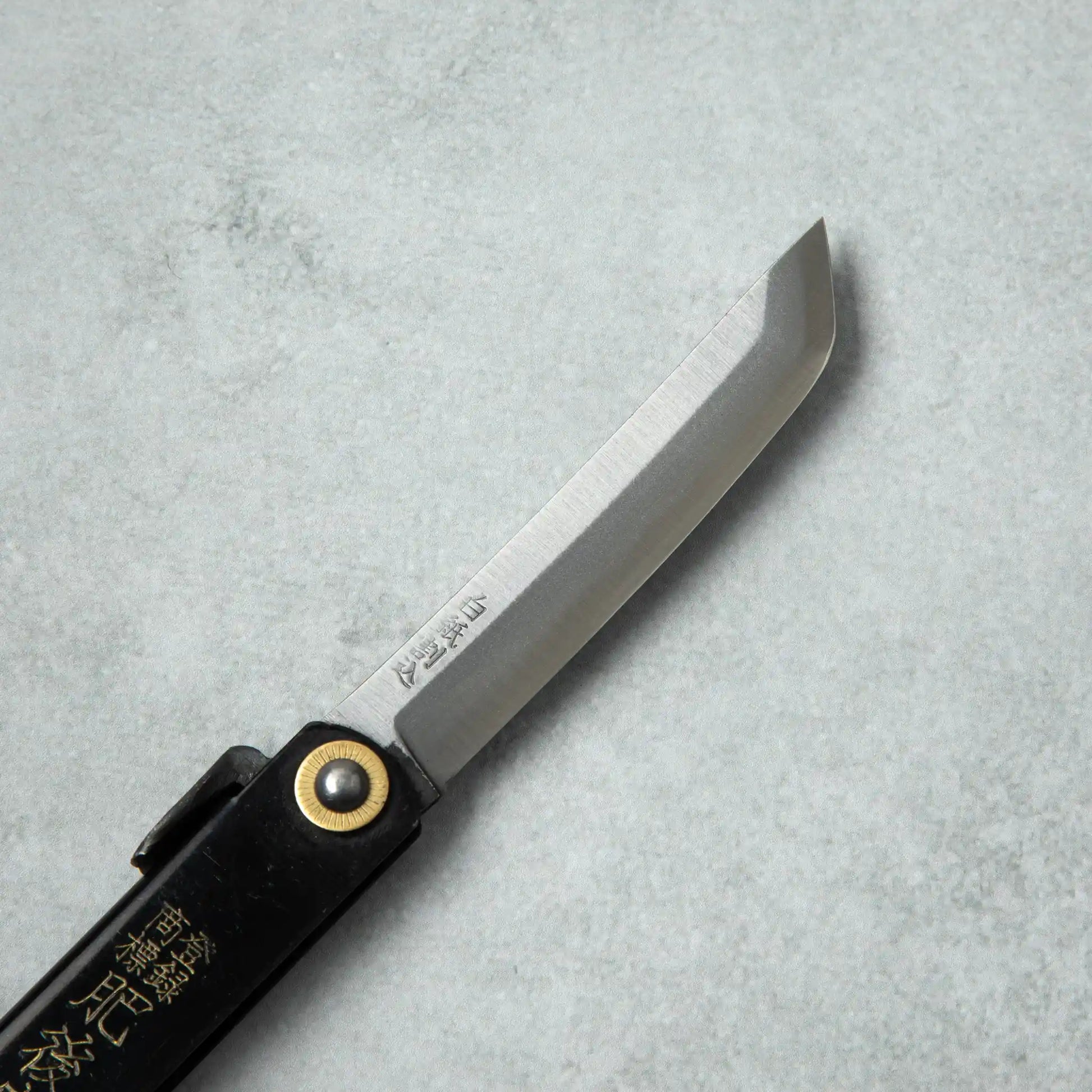 Side view of the blade of a Higonokami White Steel Sword knife, displaying its sharp edge and traditional brass pivot attached to a black handle.