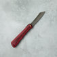 Full view of the Higonokami Woody VG10 knife with a red wooden handle and sleek, sharp blade, laid flat on a gray surface.