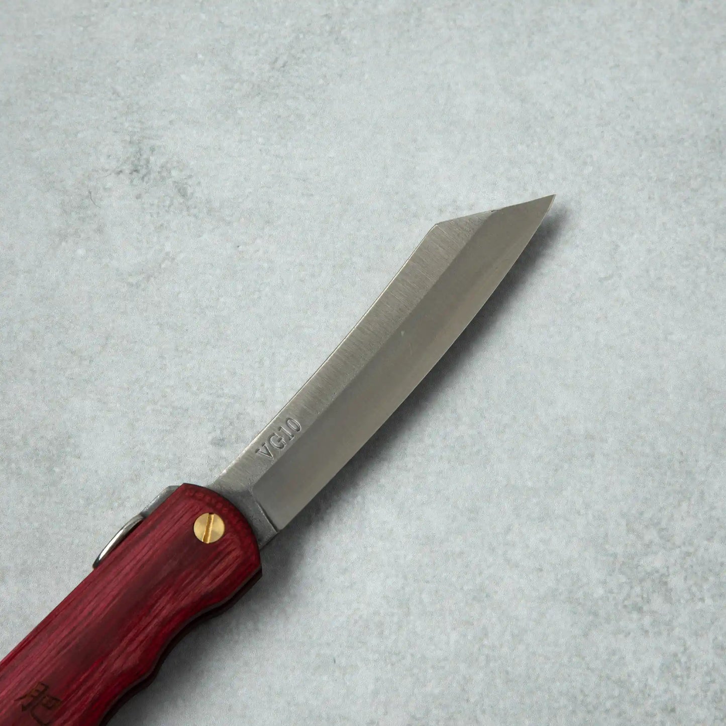 Side view of the blade of the Higonokami Woody VG10 knife, featuring the VG10 marking and brass pivot, attached to the red wooden handle.