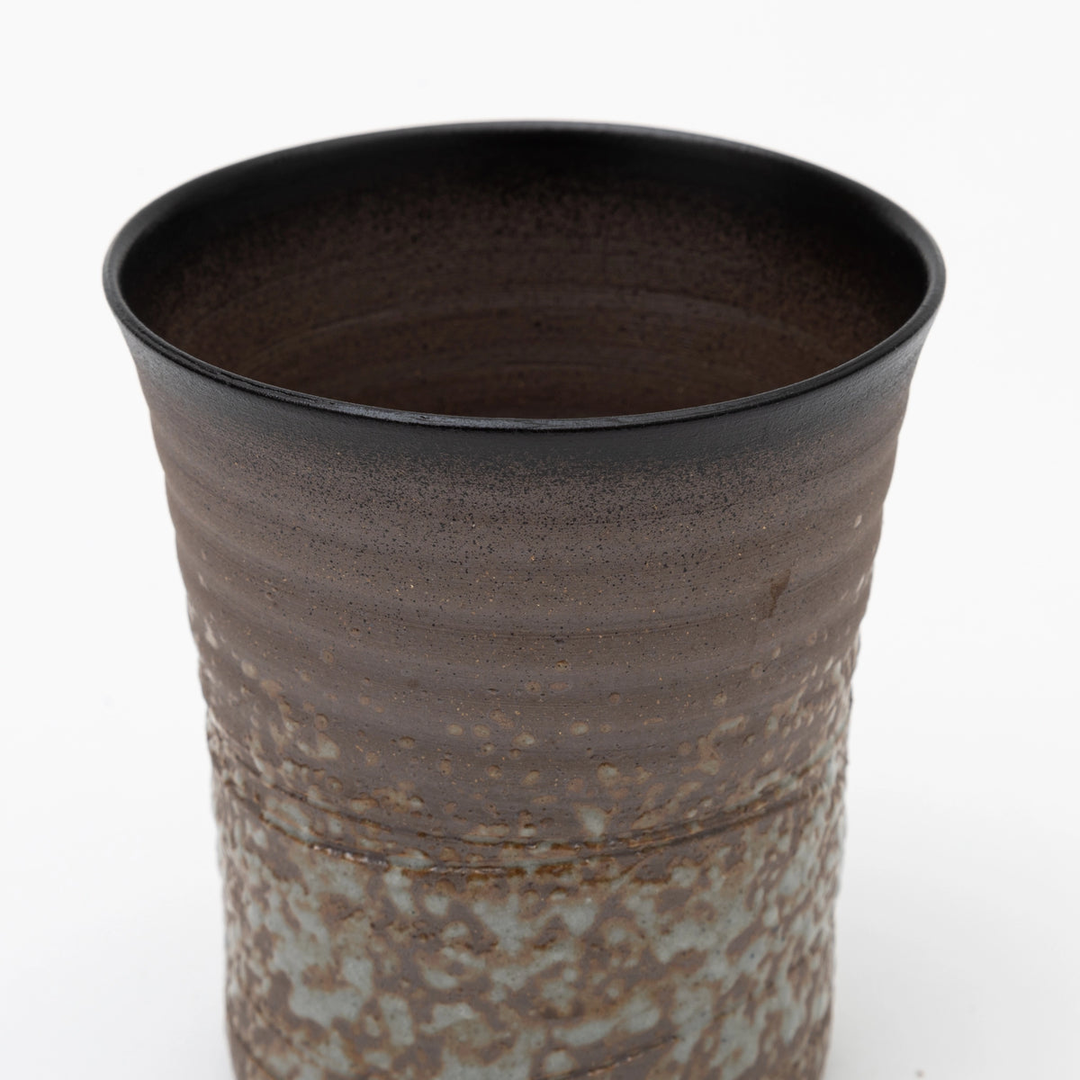 A close up of the upper part of a handleless ceramic tumbler featuring a dark brown ribbed upper section and a heavily textured, speckled grey and brown glaze on the lower half, set against a white background.
