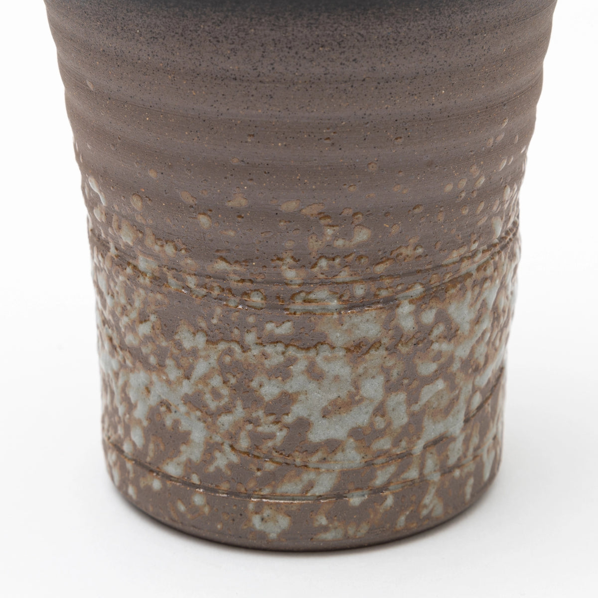 A close up of the lower part of a handleless ceramic tumbler featuring a dark brown ribbed upper section and a heavily textured, speckled grey and brown glaze on the lower half, set against a white background.
