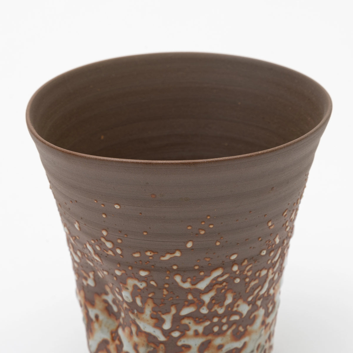 A close up of the upper part of a handleless brown ceramic tumbler featuring a  heavily textured, speckled white glaze on the lower half, set against a white background.