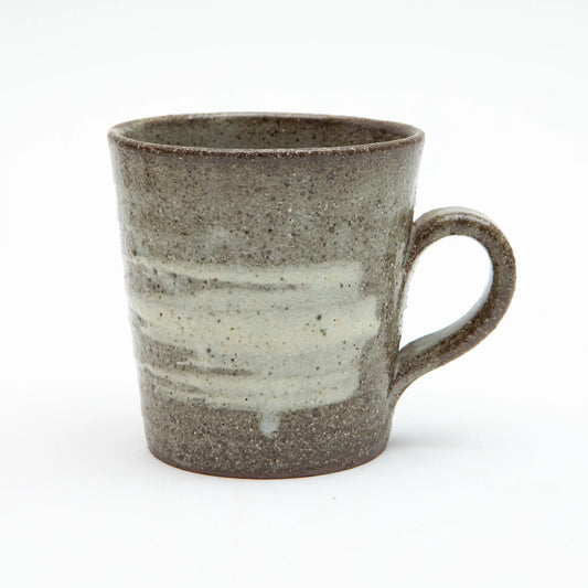 A large Shigaraki ware mug by Hinomigama with a speckled dark grey ash glaze and broad horizontal white hakeme brushstroke slip across the body, with a hand formed loop handle, set against a white background.