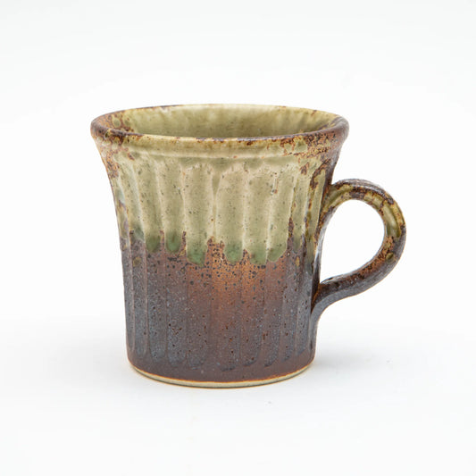 A Shigaraki ware mug by Hinomigama with vertical carved grooves, a deep charred iron brown and dark grey lower body, and a pale celadon ash glaze trailing from the rim, set against a white background.