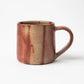 A side view of the Hozan Kiln Hidasuki Mug, displaying a warm earthy gradient of red and beige tones with natural streaks.