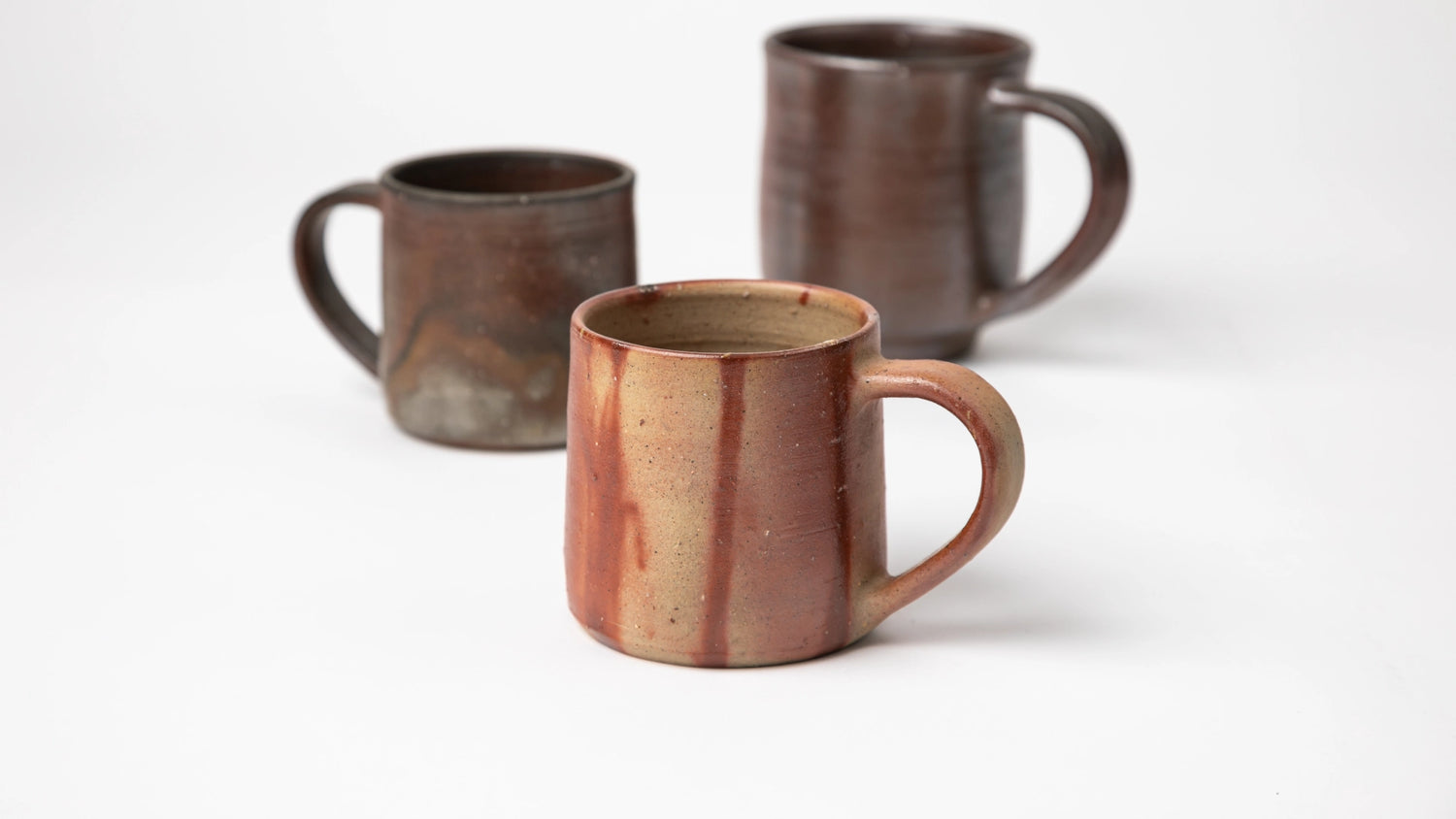 A group shot of Hozan Kiln mugs, highlighting their individual patterns and finishes with one beige and red mug in the foreground, and two dark-toned mugs in the background.