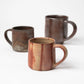 Three handcrafted mugs by Hozan Kiln displayed together, featuring unique glazing techniques in earthy red and dark tones against a white background.