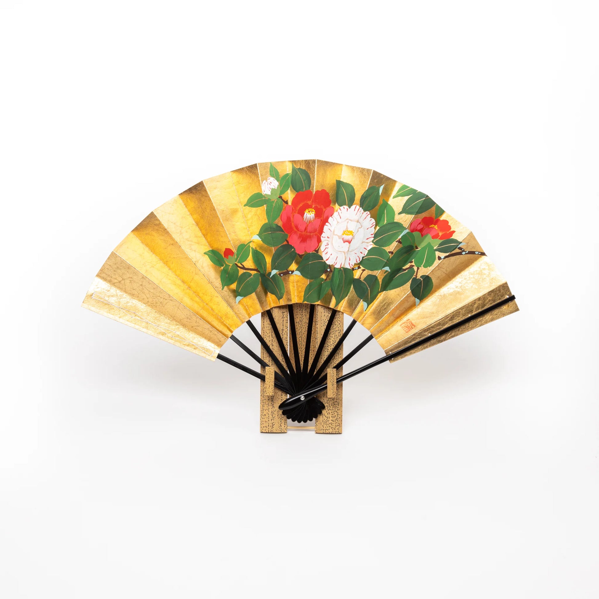 A traditional Japanese folding fan with a vibrant gold foil background, featuring hand-painted camellia flowers in vivid red and white hues, displayed on a wooden stand.