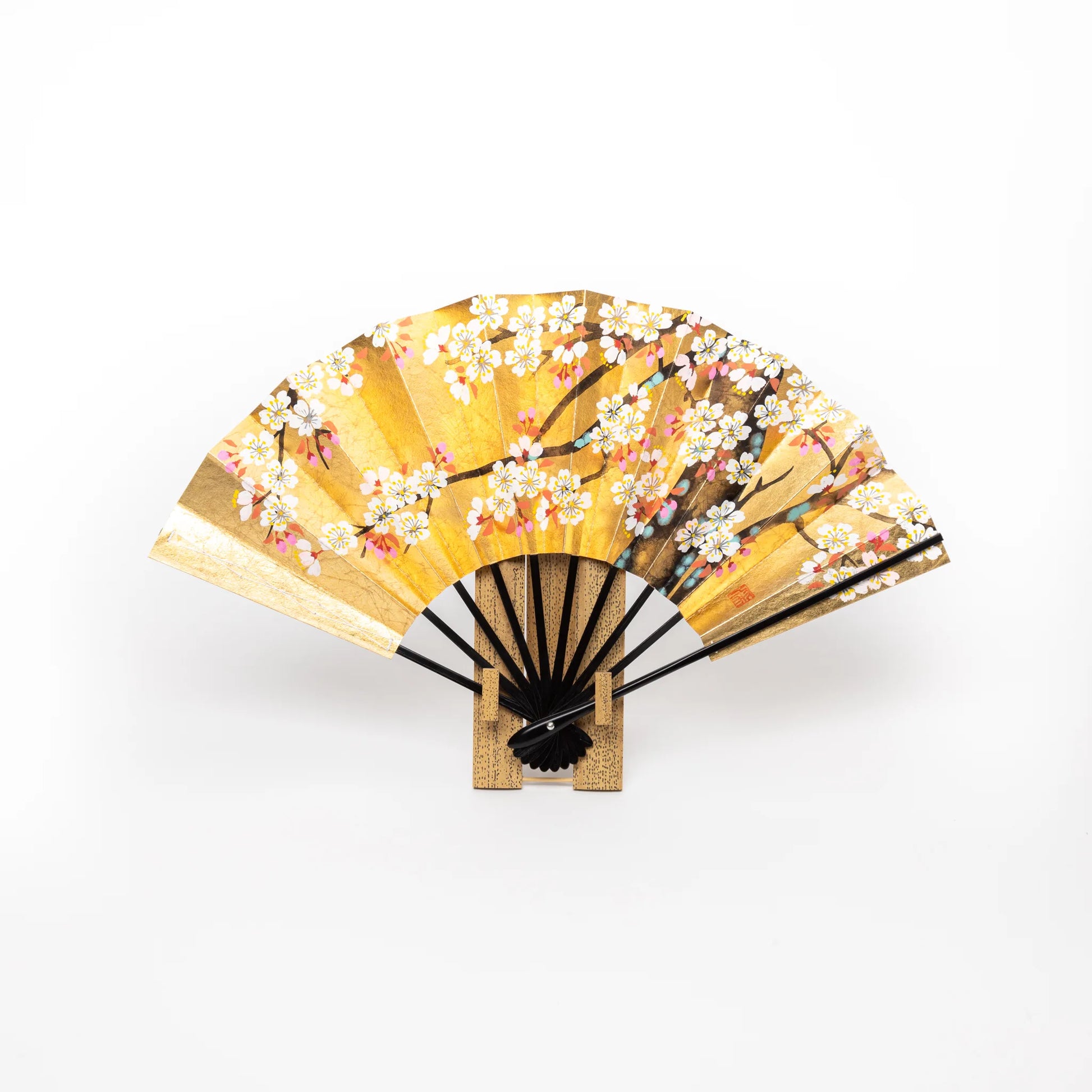 A traditional Japanese folding fan adorned with a gold background, showcasing hand-painted cherry blossom branches in full bloom, delicately presented on a wooden stand.