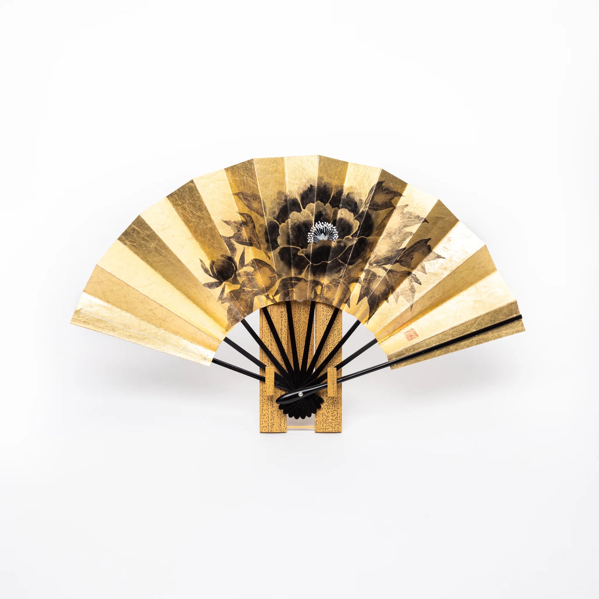 A Japanese folding fan with a luxurious gold leaf background, adorned with a black peony motif, displayed upright on a wooden stand.