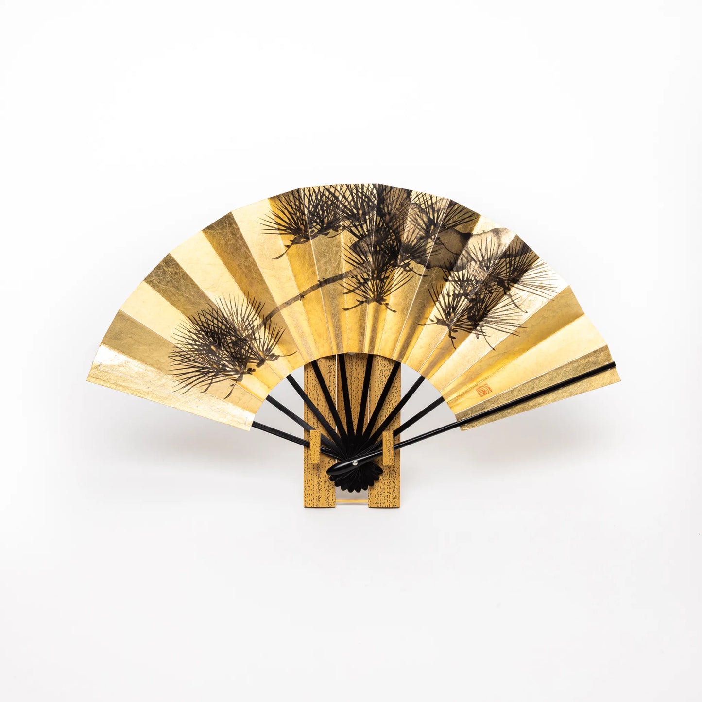 A Japanese folding fan featuring a black pine tree design on a rich gold leaf background, elegantly mounted on a display stand.