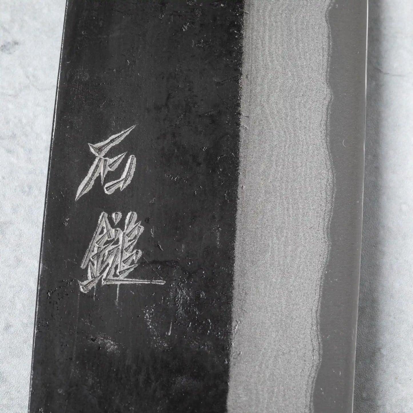 Detailed view of the kanji engraving on the Damascus-patterned blade of the Ishizuchi Blue Steel No.2 Santoku Knife, emphasizing traditional Japanese artistry.