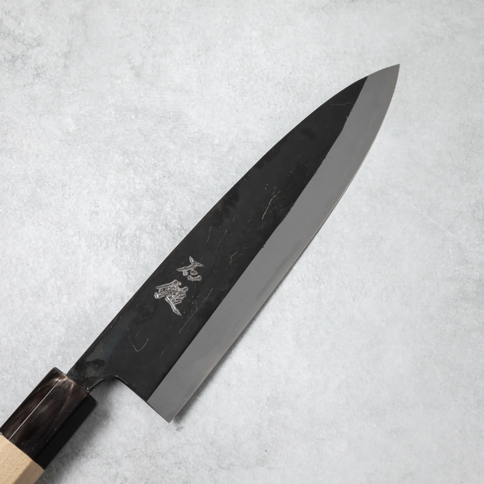 Close-up of the blade on the Ishizuchi Blue Steel No.2 Chef Knife with engraved kanji characters on a light background.