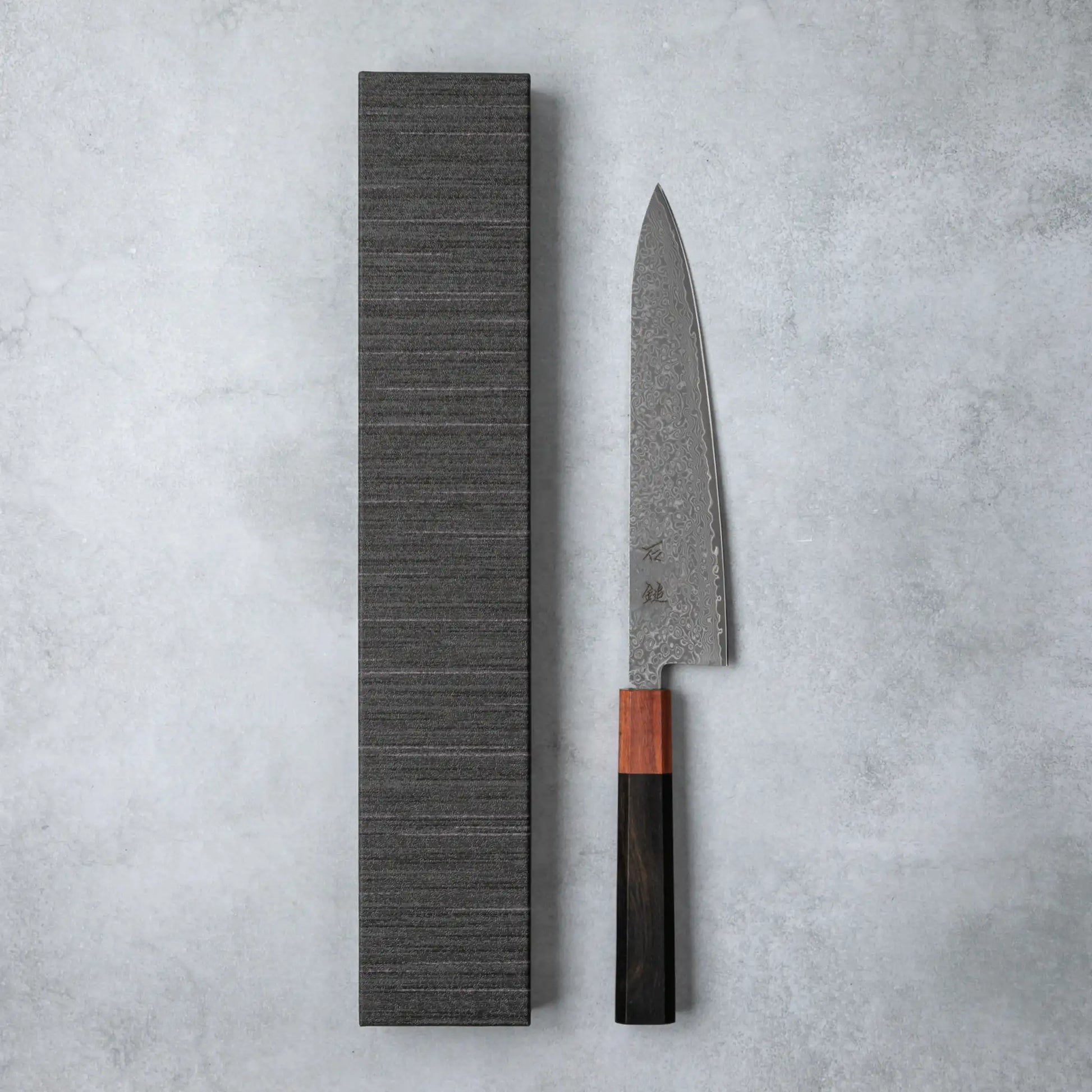 Ishizuchi VG10 Damascus Chef Knife with a polished ebony wood handle displayed alongside its textured presentation box on a gray stone surface.