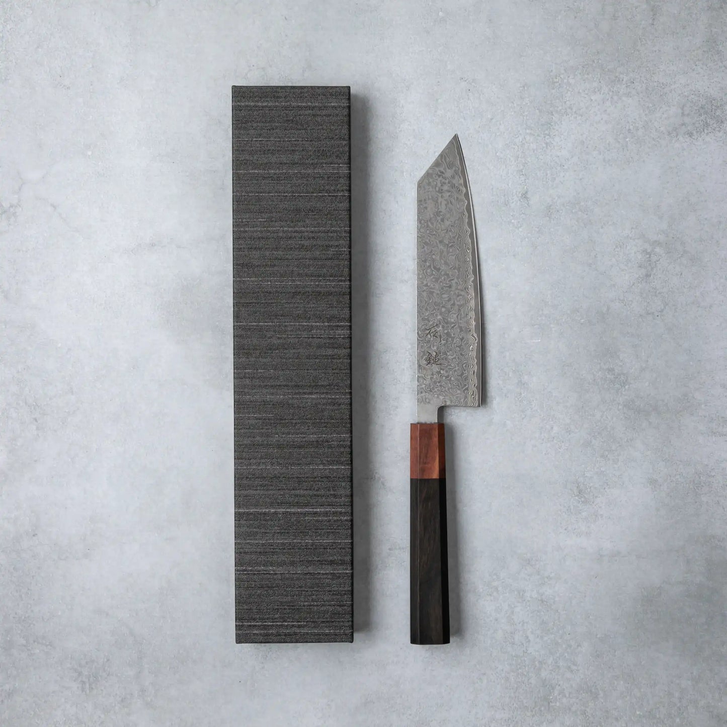 Ishizuchi VG10 Damascus Kiritsuke Knife with a polished ebony wood handle displayed alongside its elegant textured presentation box on a minimalist gray surface.