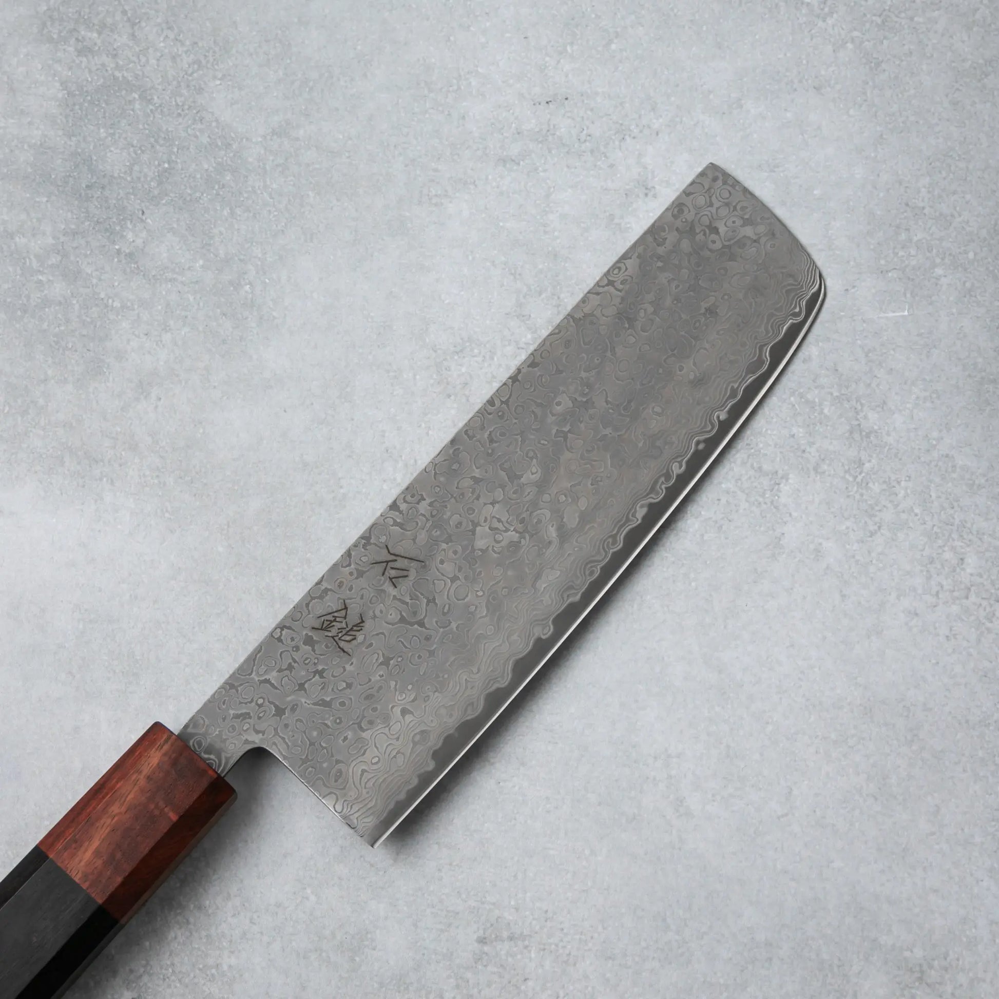 Close-up of the Damascus steel blade on the Ishizuchi VG10 Nakiri Knife, showcasing its intricate wave-like patterns and kanji engravings.