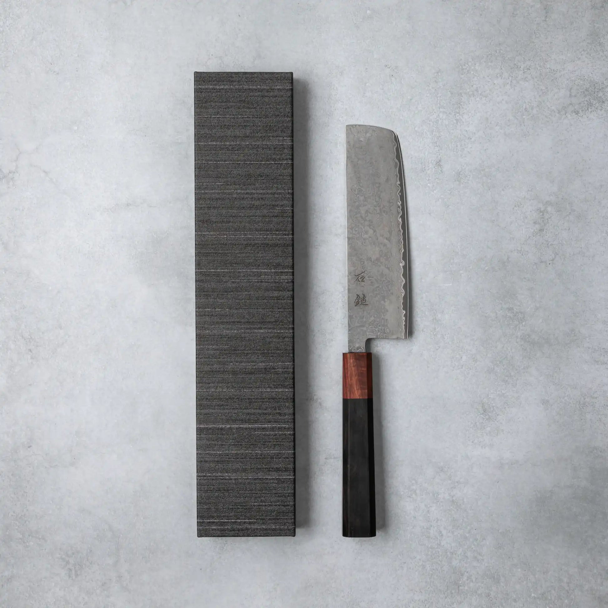 Ishizuchi VG10 Damascus Nakiri Knife with a refined ebony wood handle displayed alongside its textured gray presentation box on a minimalist gray background.