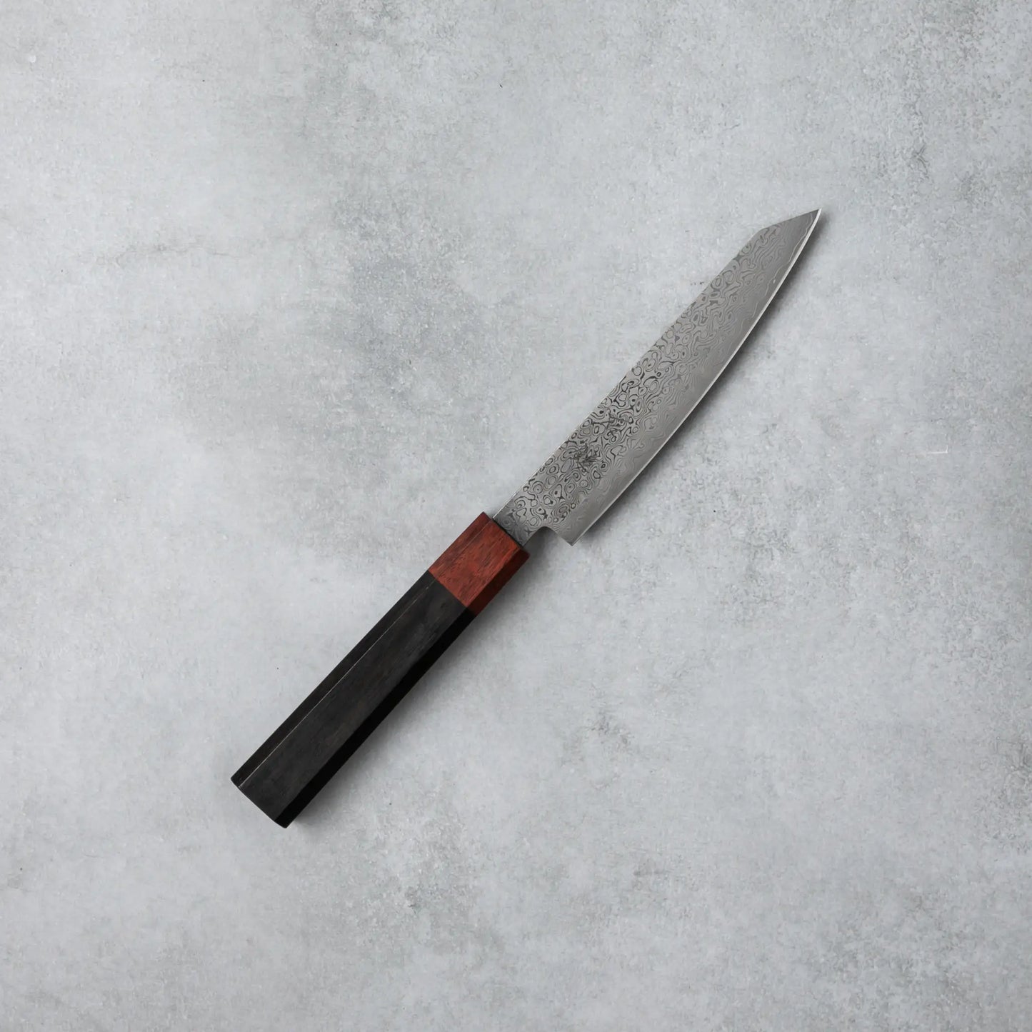 Full view of the Ishizuchi VG10 Damascus Petty Knife, showcasing its compact, sharp blade and refined ebony wood handle, set against a plain gray surface.