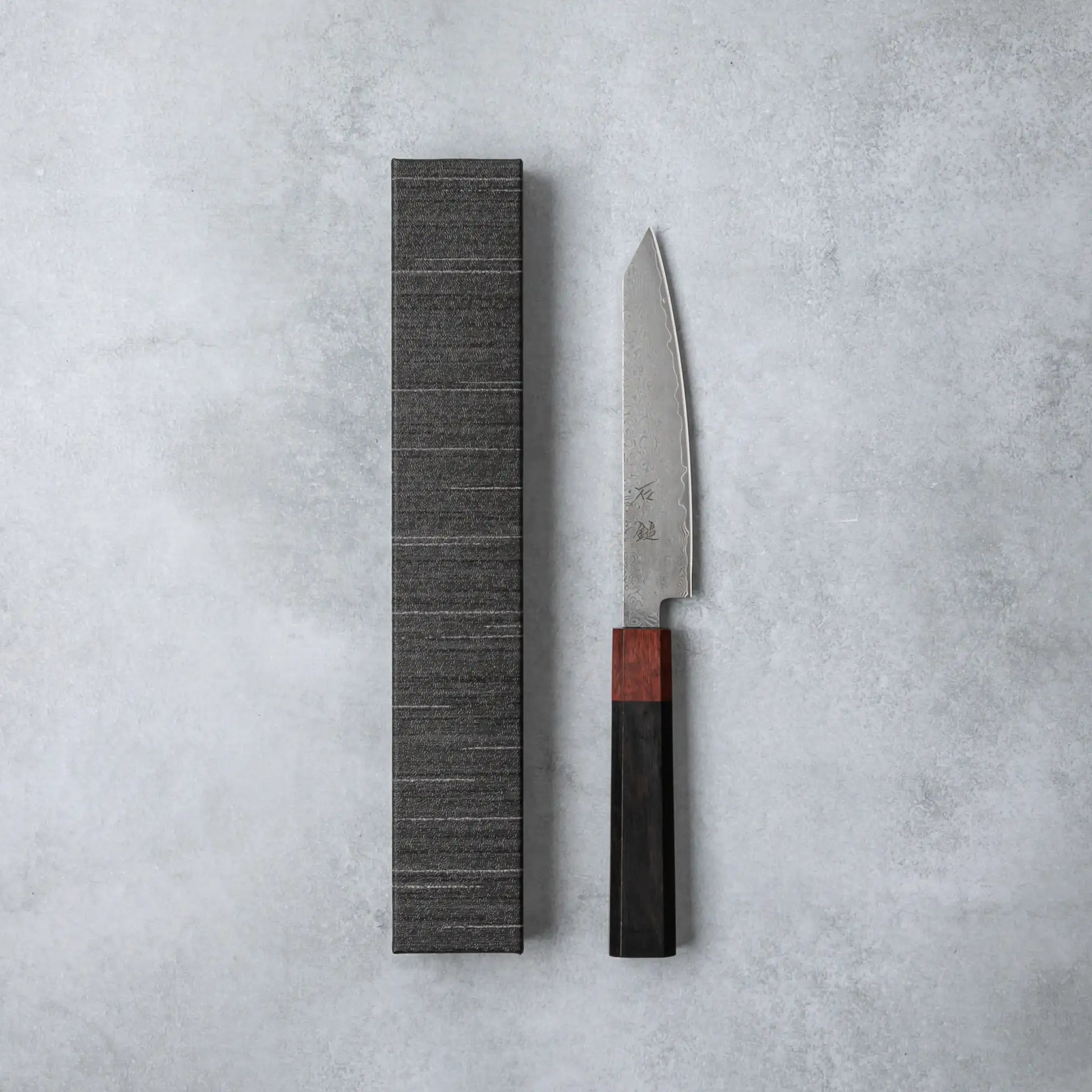 Ishizuchi VG10 Damascus Petty Knife with a polished ebony wood handle displayed alongside its textured gray presentation box on a minimalist gray background.