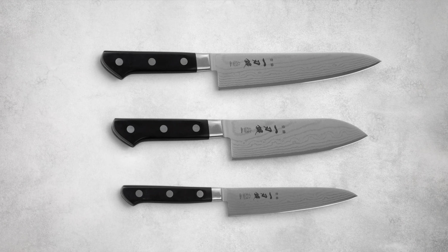 A set of three Ittoutan Kiageha knives arranged in a horizontal layout on a textured stone surface. The set includes a chef’s knife, a Santoku knife, and a petty knife, all featuring Damascus steel blades and black handles with triple-rivet construction.