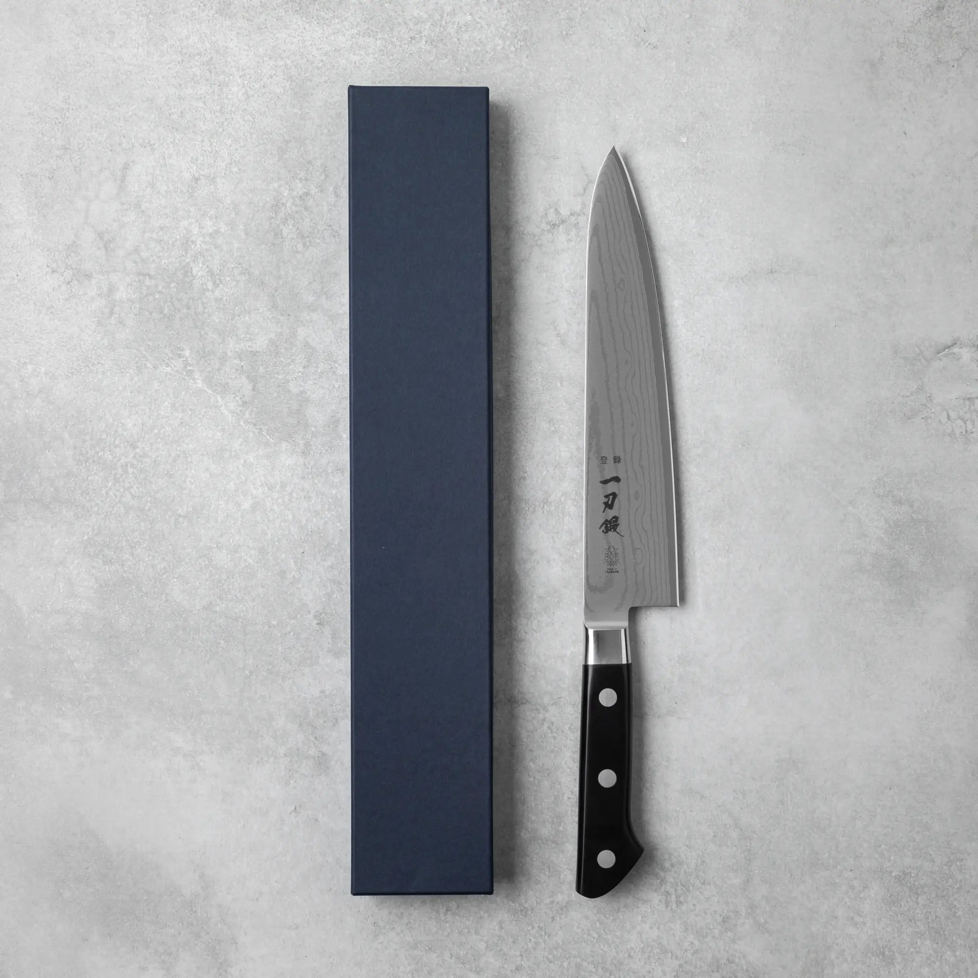 A Japanese chef’s knife with a Damascus steel blade, displayed beside its sophisticated navy blue packaging.