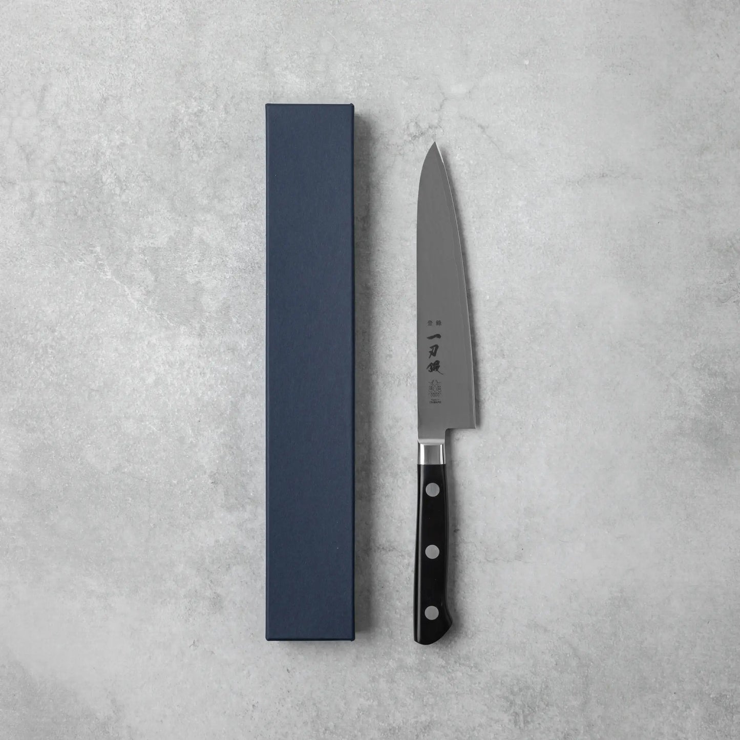 A Japanese petty knife with a Damascus steel blade and black handle, placed beside a navy-blue box on a textured gray surface.