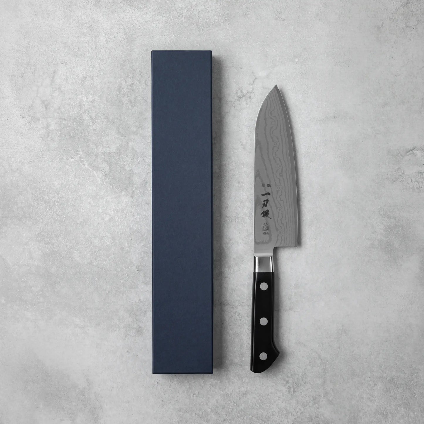 A Japanese Santoku knife with a Damascus steel blade and black handle, placed beside a navy blue box on a grey stone surface.