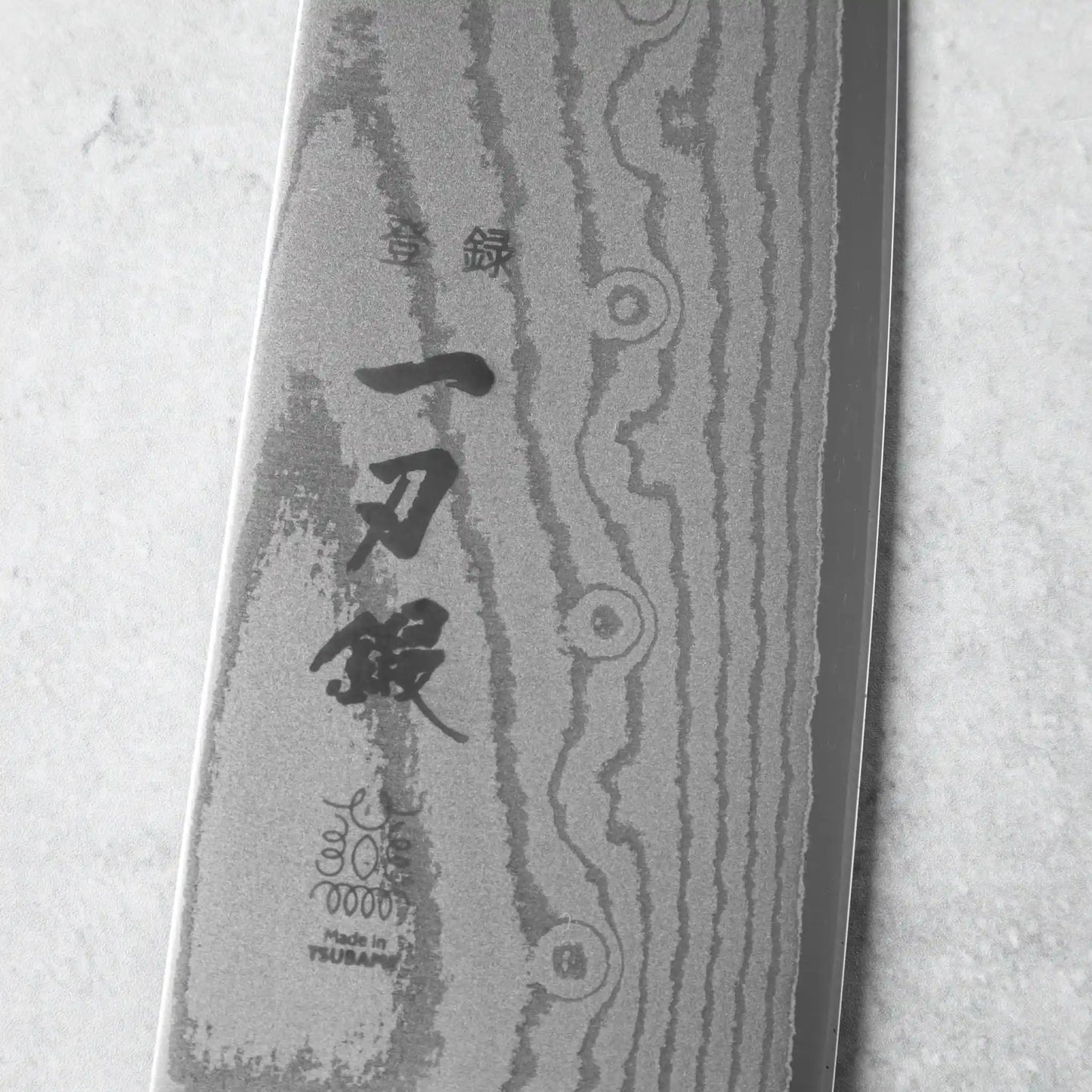 The engraved Japanese characters and branding on the Ittoutan Kiageha Santoku knife’s Damascus steel blade.