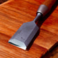 A close-up of a single Kakuri Damascus chisel resting on a wooden surface, showcasing the intricate Damascus steel pattern on the blade.