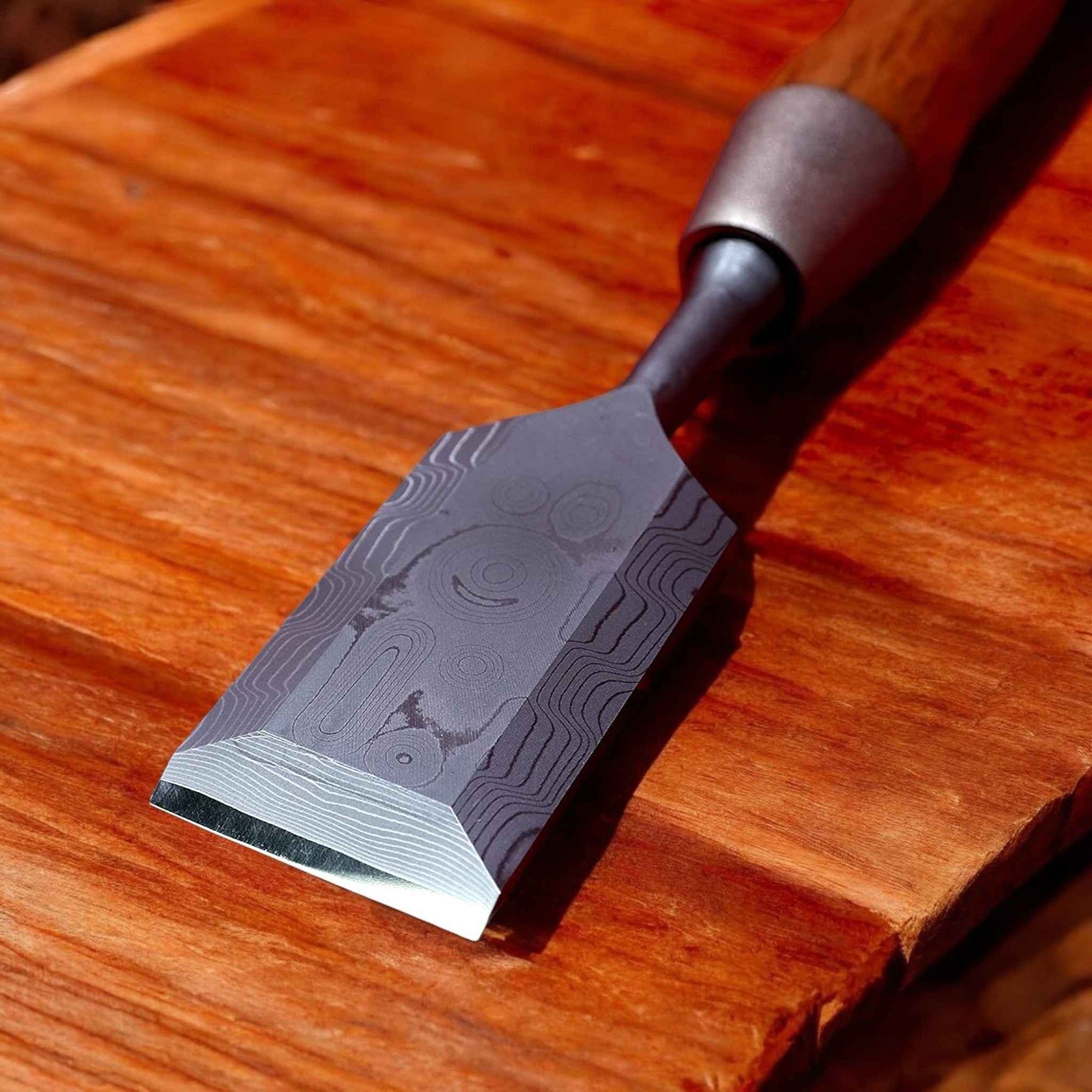 A close-up of a single Kakuri Damascus chisel resting on a wooden surface, showcasing the intricate Damascus steel pattern on the blade.