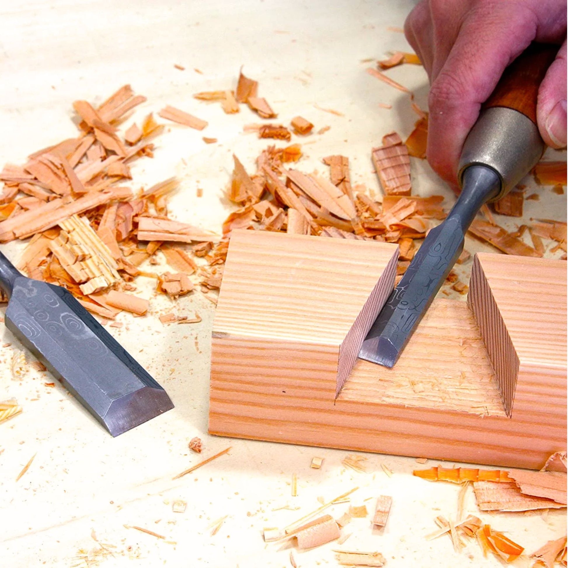 A Kakuri Damascus chisel in action, cutting a precise groove into a piece of light-colored wood, with wood chips scattered around.