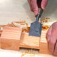 A close-up of a woodworker's hands using a Kakuri Damascus chisel to carve a joint into a piece of wood, surrounded by wood shavings.