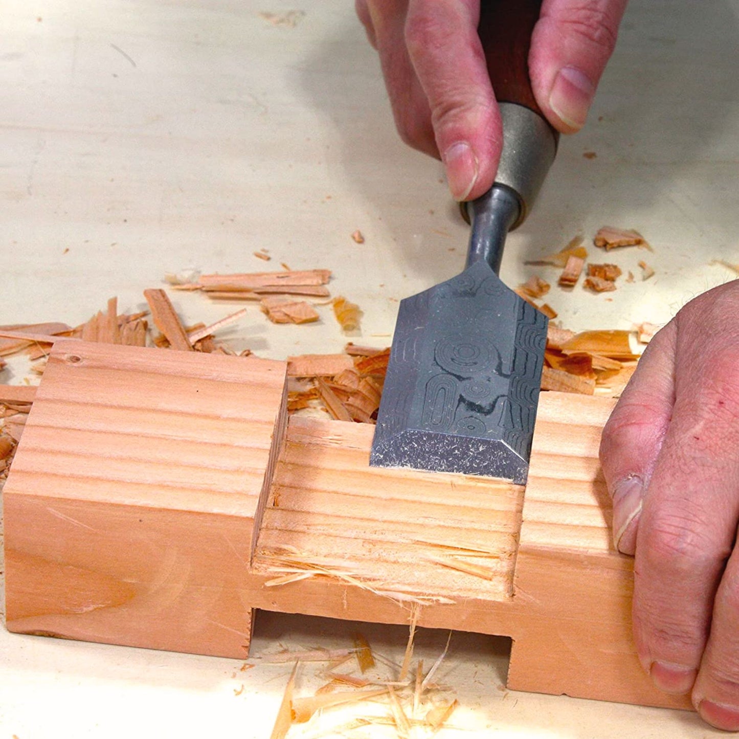 A close-up of a woodworker's hands using a Kakuri Damascus chisel to carve a joint into a piece of wood, surrounded by wood shavings.