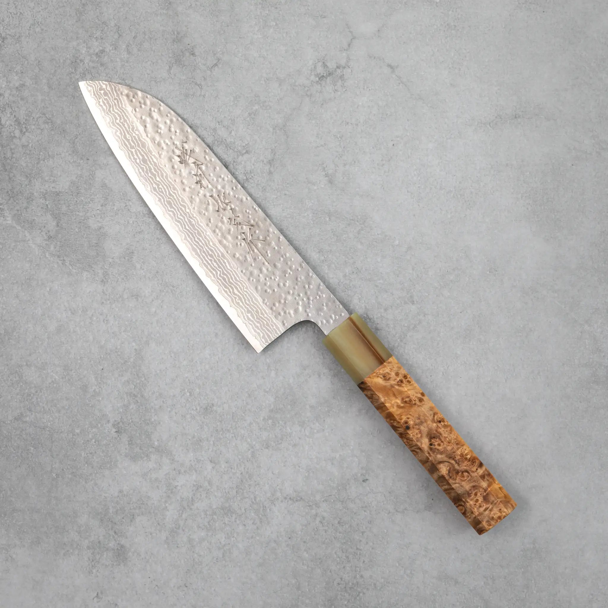 Side view of the Kamo Hamono Shizuku santoku knife, showing a VG10 Damascus blade with rounded tsuchime hammer marks across the upper surface, a water buffalo horn bolster, and a stabilized burl wood handle with pronounced figured grain.You said: stabilized burr wood?
