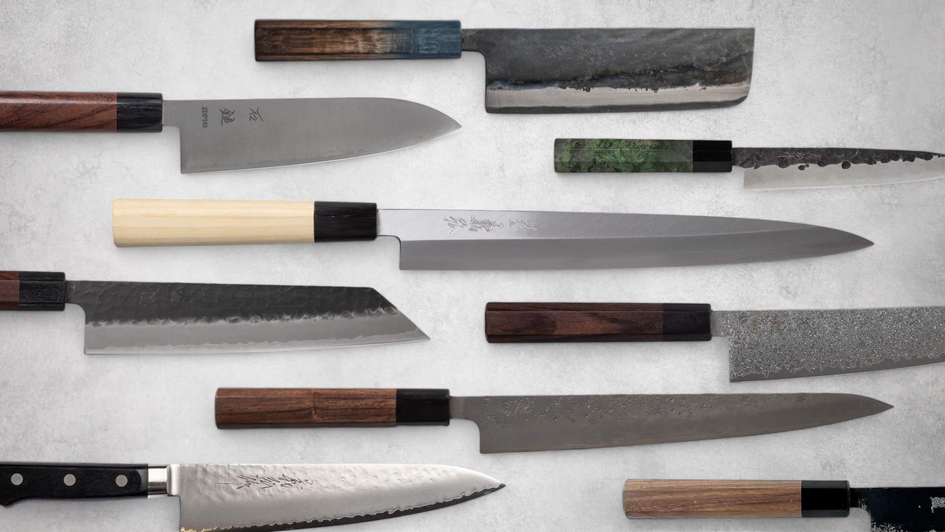 Assorted Japanese knives with a variety of blade styles and handles arranged on a light surface. The collection features traditional craftsmanship with different materials, blade finishes, and wooden handles, showcasing the diversity and artistry of Japanese knife-making.